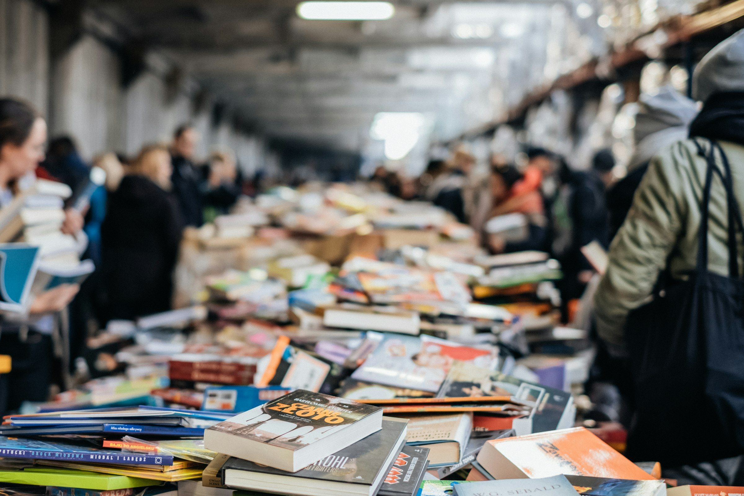 Mesas de uma feira do livro cobertas de livros, com o corredor cheio de visitantes ao fundo.